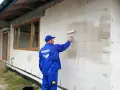 Pracownik w niebieskim kombinezonie nakłada wałkiem grunt na ścianę z bloczków. To kluczowy etap przygotowania pod styropian.