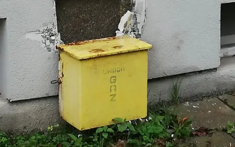 Żółta skrzynka gazowa w elewacji budynku, z napisem "UWAGA GAZ".