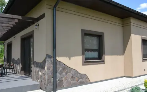 Elewacja domu w kolorze kawa z mlekiem, ozdobiona kamiennym cokołem. Widoczne okna i drzwi tarasowe.