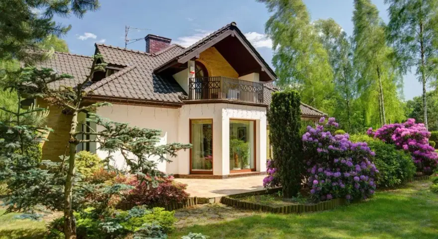 Dom z balkonem i tarasem, otoczony bujną zielenią i kwitnącymi rododendronami. Występ ściany podkreśla nowoczesny charakter budynku.