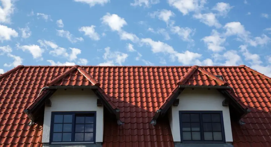 Czerwony dach z dachówki ceramicznej z dwoma lukarnami na tle błękitnego nieba z chmurami.